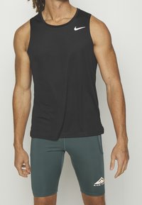 Homme portant un débardeur noir Nike et un short de trail Nike vert foncé, debout devant un fond uni.