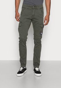Homme portant un pantalon cargo vert olive avec des patchs et des baskets noires à semelles blanches, debout devant un fond uni.