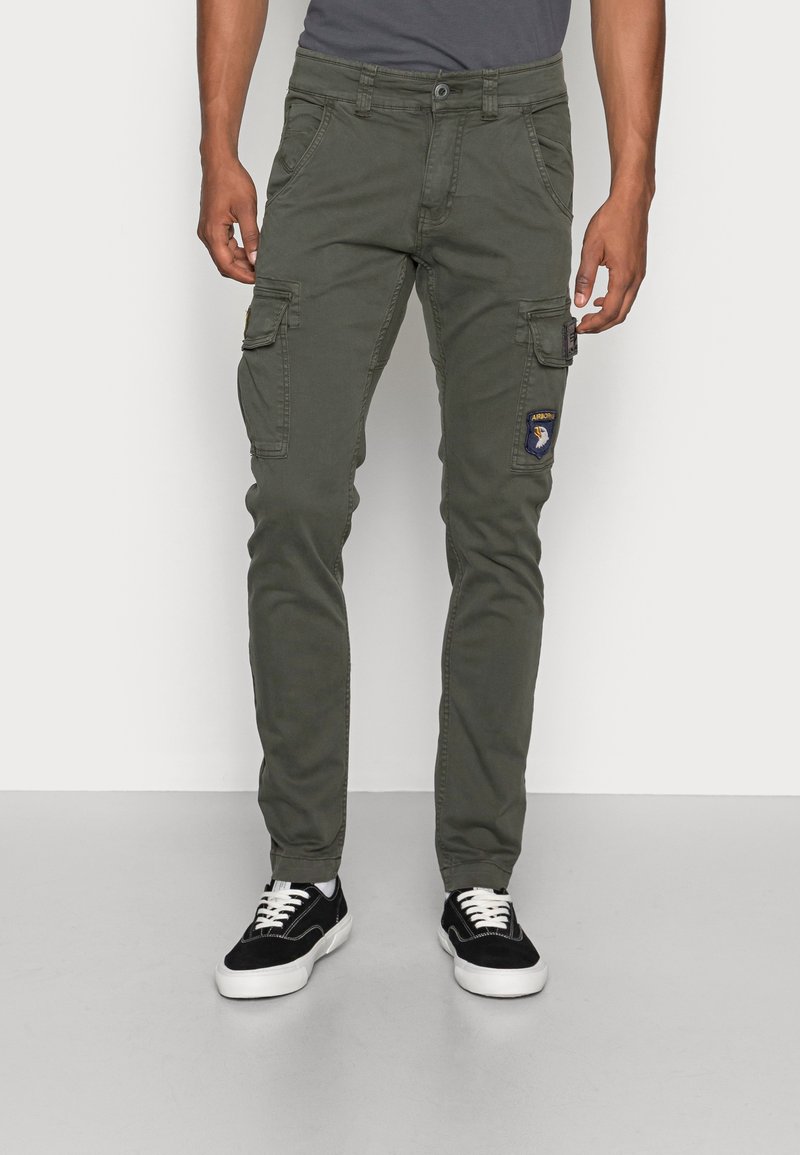 Homme portant un pantalon cargo vert olive avec des patchs et des baskets noires à semelles blanches, debout devant un fond uni.