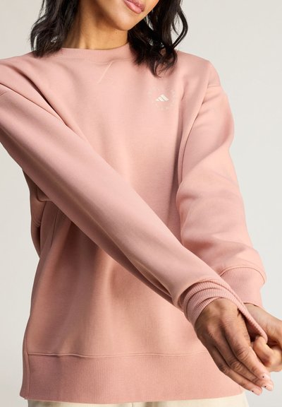 Kvinde iført en lyserød sweatshirt med lange ærmer og et lille hvidt logo nær skulderen, som justerer ærmet med hænderne.