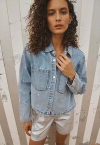 Lichtblauwe denim jas met twee voorkruiszakken, knoopsluiting, geborduurde logo's en lange mouwen, gecombineerd metlichte shorts.