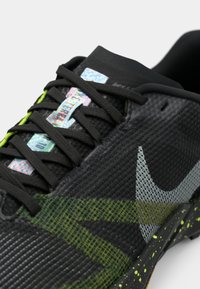 Prim-plan al unui pantof sportiv negru cu accente verzi, șireturi negre, o etichetă reflectorizantă pe limbă și un swoosh Nike texturat pe lateral.