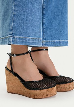 Keilsandalen mit schwarzem Mesh-Obermaterial und Knöchelriemen, getragen zu blauen, weit geschnittenen Jeans, die an den Knöcheln enden.