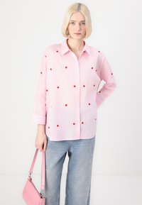 Light pink button-up shirt with embroidered red hearts, long sleeves, and a relaxed fit, paired with light blue jeans featuring heart patterns.