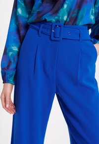 Pantalon ajusté bleu vif avec une large ceinture et une boucle carrée, présentant des plis avant et une texture lisse. Associé à un chemisier à motif multicolore.