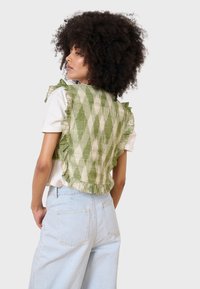 Gilet verde con motivo a righe e bordi arricciati, caratterizzato da un design a scacchi. Indossato sopra una maglietta bianca, abbinato a jeans a gamba larga blu chiaro.