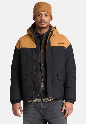 Uomo che indossa una giacca Timberland nera e marrone, berretto marrone, sopra una camicia a quadri e jeans scuri su uno sfondo semplice.