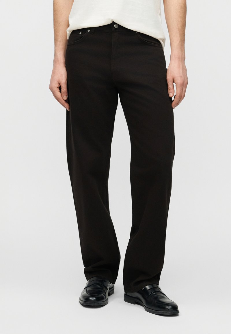 Άνδρας που φοράει φαρδιά μαύρη παντελόνα, μαύρα  δερμάτινα παπούτσια χωρίς κορδόνια και λευκό πουκάμισο, σε απλό φωτεινό φόντο.