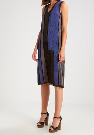 Femme portant une robe sans manches longueur genoux à rayures verticales bleues, noires et grises, avec des sandales à plateforme à talons hauts noires.