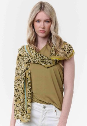 Junge Frau mit langen blonden Haaren trägt ein olivgrünes Oberteil, weiße Hosen und einen senfgelben, gemusterten Schal, der über eine Schulter drapiert ist.