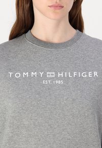 Szara bluza z okrągłym dekoltem, z wyraźnym białym napisem: "TOMMY HILFIGER EST. 1985" na przodzie. Gładka tekstura, casualowy design.