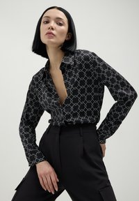 Donna con un taglio di capelli bob nero, che indossa una camicia nera a maniche lunghe con un motivo e pantaloni neri a vita alta, in posa su uno sfondo neutro.