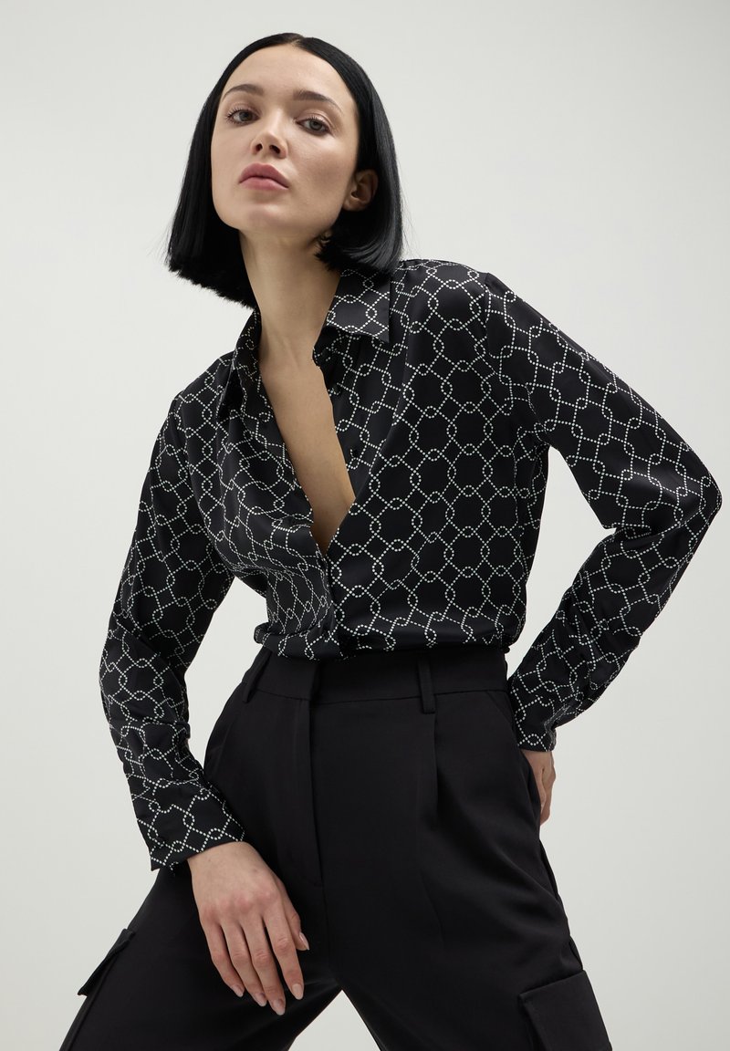 Donna con un taglio di capelli bob nero, che indossa una camicia nera a maniche lunghe con un motivo e pantaloni neri a vita alta, in posa su uno sfondo neutro.