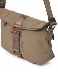 Beige schoudertas met een overslagklep, verstelbare bruine band, metalen beslag en een oranje gesp. Heeft een logo dat op de zijkant is gedrukt.