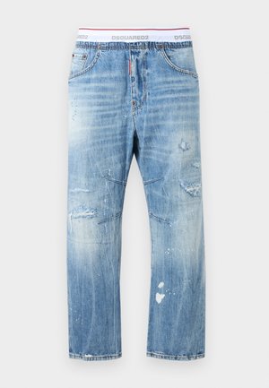 Νέος άνδρας που φοράει λευκό φανελάκι, ανοιχτό μπλε ταλαιπωρημένο τζιν, πράσινα αθλητικά παπούτσια και ανοιχτό μπλε καπέλο με την επιγραφή "ROOT".