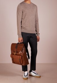 Zaino in pelle marrone con pattina, tasca frontale e accessori in oro, abbinato a pantaloni neri e sneakers bianche-grigie su uno sfondo neutro.