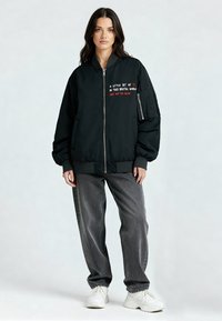 Zwarte bomberjack met een ritssluiting aan de voorkant, voorzien van geborduurde tekst op de borst en een zak op de linkerarm. Gecombineerd met losse grijze spijkerbroeken en witte sneakers.