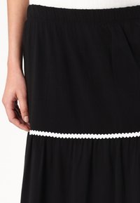 Jupe noire avec un design froncé, une taille élastique et un ourlet orné d'une dentelle blanche. La texture du tissu doux est visible.