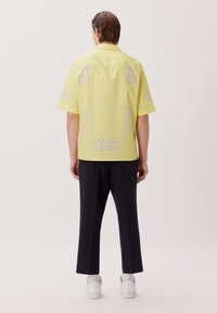 Camicetta gialla a maniche corte con motivo geometrico bianco, indossata con pantaloni neri e sneakers bianche, vista da dietro.