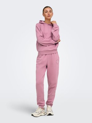 Jeune femme debout, les bras croisés, portant un sweat à capuche rose assorti et un pantalon de jogging avec des baskets blanches, sur un fond clair uni.