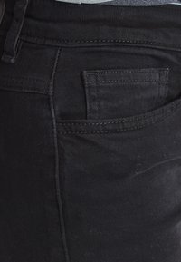 Los jeans de mezclilla negra tienen un diseño ajustado con un pequeño bolsillo trasero. La tela tiene una textura suave y un sutil detalle de costura.