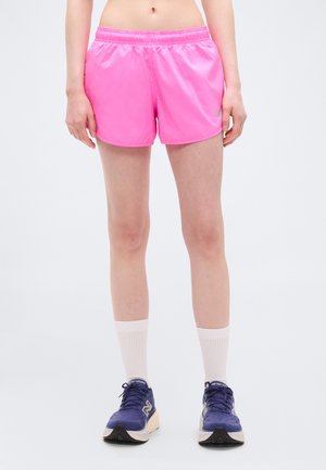 Frau trägt leuchtend pinke Sportshorts, weiße Sportsocken und blaue Laufschuhe, steht vor einem schlichten hellen Hintergrund.