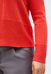 Άτομο που φοράει κόκκινο πλεκτό πουλόβερ με μακριά μανίκια σε συνδυασμό με γκρι παντελόνι, με το χέρι χαλαρό δίπλα στο σώμα, μπροστά σε απλό φόντο.