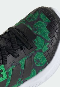 Zapatilla deportiva negra con un diseño gráfico pixelado en verde, parte superior de malla texturizada, detalles sintéticos negros y suela de goma blanca.