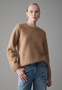 Lysebrun, fuzzy strikkegenser med rund hals og lange ermer, kombinert med lyseblå denimjeans i en avslappet passform.