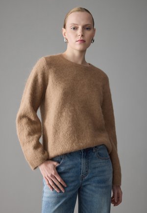 Lysebrun, fuzzy strikkegenser med rund hals og lange ermer, kombinert med lyseblå denimjeans i en avslappet passform.