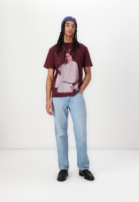 Maroon t-shirt featuring a graphic print of a woman in a grey outfit. Worn with light blue jeans and black shoes. Blue knit beanie on head.