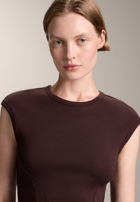 Burgunderfärgad ribbad ärmlös topp med en figurnära passform och rundad halsringning. Slät textur med böjda sidokanter för en figursydd struktur.