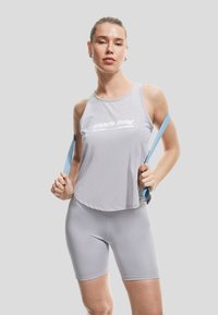 Hellgraues Sporttanktop mit weißem Text und passenden Radshorts. Glattes, leichtes Material. Das Modell hält hellblaue Träger.