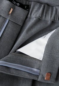 Pantalons en tissu gris avec une ceinture élastique, équipés d'une poche zippée et d'une doublure intérieure blanche. Accentué par un petit détail en métal.