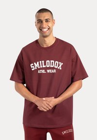 Bordeaux oversized t-shirt van katoen, met een witte gebogen tekstprint "SMILODOX ATHL. WEAR" op de voorkant. Zachte, gladde textuur.