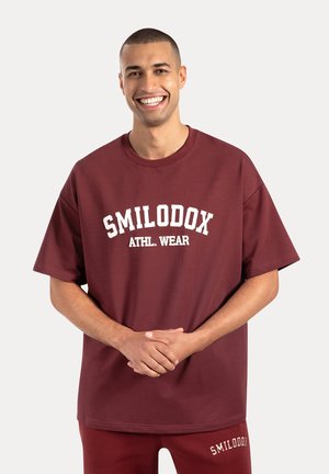 Bordeaux oversized t-shirt van katoen, met een witte gebogen tekstprint "SMILODOX ATHL. WEAR" op de voorkant. Zachte, gladde textuur.