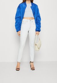 Chaqueta bomber satinada azul, top corto de un azul claro, jeans skinny blancos desgastados, tacones negros con tiras y un bolso de hombro color crema.