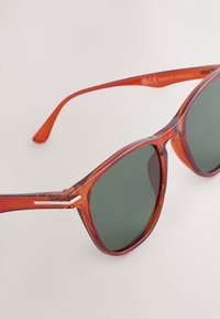 Oranje doorschijnende zonnebril met donker groene lenzen, close-up weergegeven op een effen lichte achtergrond.