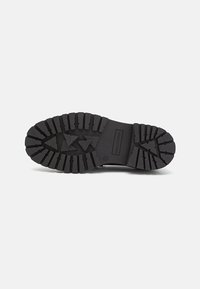 Semelle de chaussure noire avec un profond motif de bande de roulement, présentant des rainures triangulaires et rectangulaires pour l'adhérence, montrée sur un fond clair.