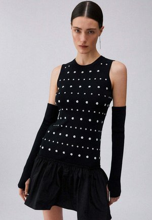 Mujer con el pelo peinado hacia atrás lleva un vestido negro sin mangas con lunares blancos, calentadores de brazos negros y una minifalda negra con volantes.