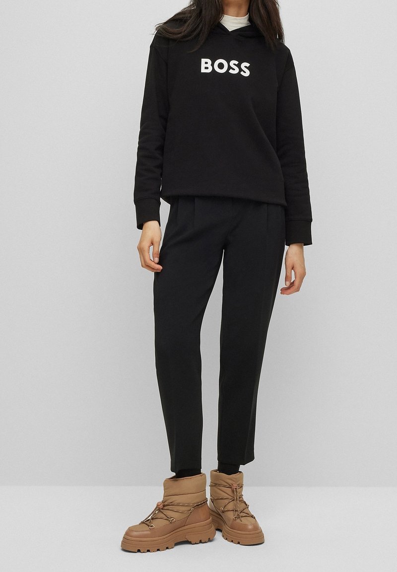 Femme portant un sweat-shirt noir "BOSS", un pantalon noir et des bottes d'hiver brunes à lacets, debout devant un fond gris clair uni.