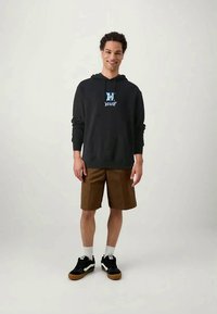 Ung man som ler, iklädd en svart hoodie med blå logotyp, bruna shorts, vita strumpor och svarta sneakers, stående mot en enkel ljus bakgrund.