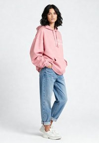 Felpa oversize rosa con testo bianco, abbinata a jeans azzurri arrotolati e sneakers bianche, in piedi davanti a uno sfondo chiaro.