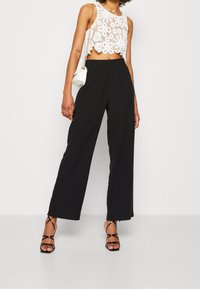Bílý háčkovaný crop top s květinovým vzorem, zkombinovaný s černými kalhotami s širokými nohavicemi. Černé sandály na vysokém podpatku a malá bílá kabelka dotváří celkový vzhled.