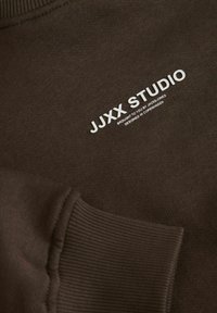 Bruine sweatshirt met ribgebreide mouwen en halslijn. Bevat witte tekstbranding: "JJXX STUDIO" en extra kleine tekst eronder.