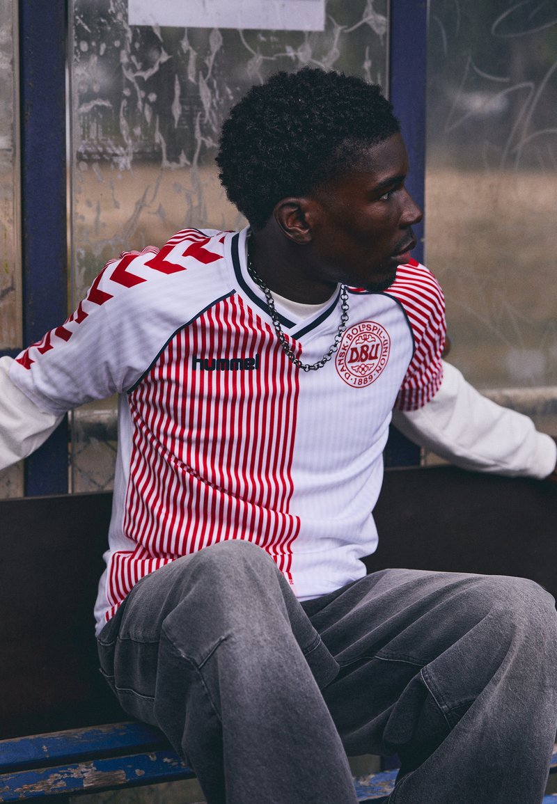 White football jersey with red vertical stripes, black branding, and a circular logo. Paired with loose grey pants.