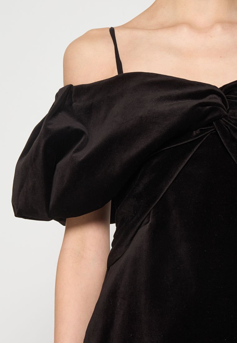 Robe en velours noir avec un design décolleté. Elle présente un devant noué et des manches bouffantes structurées. Fines bretelles.