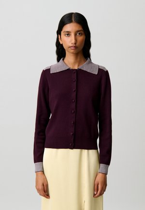 Donna che indossa un cardigan bottonato viola scuro con collo e polsini a righe, abbinato a una gonna beige chiaro, in piedi davanti a uno sfondo semplice.