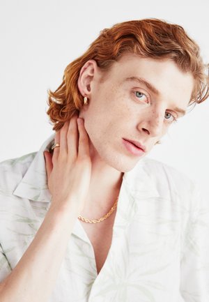 Fehér, rövid ujjú ing zöld levélmintával, arany lánccal és fülbevalóval. A modellen göndör vörös haj és világos bőr található.