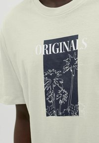 Lichtgroene katoenen t-shirt met een marineblauw rechthoekig grafisch ontwerp met witte lijngetekende palmbomen en het woord "ORIGINALS" erboven gedrukt.